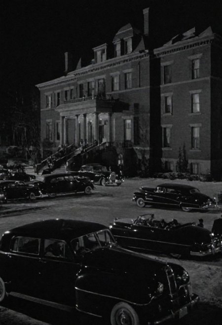 In the 1950s in Chicago, outside at night, from afar you can see a luxurious mansion of the mafia boss, you can see several cars and many armed gangsters protecting. Captured with a 35 mm camera, black and white, classic film noir film.