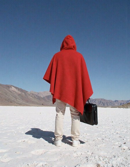 A photograph from behind, back of a person in a bright red mantle, mantle is very baggy, huge hanging sleeves, standing in a completely white desert, white sand, the sky is smooth blue. holding a black briefcase.