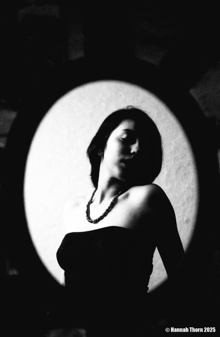 A black and white photograph from a low angle, focusing on a woman standing in the middle of the frame. the woman, who appears to be in her early twenties, has long, dark hair and is facing away from the camera, with her eyes closed. she is wearing a strapless black top that accentuates her bare shoulders and medium-sized breasts. the top is adorned with a beaded necklace that adds a touch of sparkle to the otherwise monochromatic image. the background is dark and out of focus, creating a sense of depth and dimension. the lighting is soft and diffused, casting gentle shadows that accentuate the woman's figure. the overall mood of the image is somber and contemplative, as if the woman is in a state of deep contemplation.<lora:_Flux\Saul_Leiter_BW.safetensors:1.0:1.0>