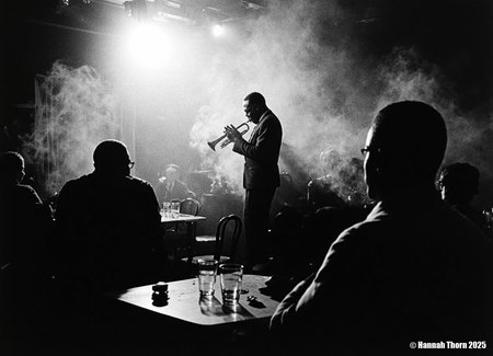The image is a black and white photograph of Miles Davis playing his trumpet.  The photo is taken in a dimly lit, smoky environment, possibly a jazz club. Davis is centrally positioned, but slightly off-center, his body angled towards the trumpet. He's captured mid-performance, his face partially obscured by shadows and the smoke, creating a dramatic and mysterious atmosphere.  He's wearing a dark suit. His focus is entirely on his instrument.  In the foreground, the backs of other patrons are visible, partially silhouetted, seated at a table with glasses, suggesting the scene is from a live performance.  The smoke adds depth and texture to the photo, and the lighting creates strong contrast, with bright spots highlighting Davis and his trumpet, and leaving the rest in shadows.  The overall feeling is intimate and moody. The photograph's style is strongly reminiscent of classic jazz photography. It uses elements of: Chiaroscuro: The dramatic use of light and shadow to create strong contrasts and a sense of depth. High Contrast: The deep blacks and bright whites add to the intensity and emotion of the scene. Documentary style: It seemingly captures a candid moment from a real-life event, without significant staging.  While composed, the image feels unplanned, spontaneous, and captures the essence of the moment. Grainy texture: The texture of the photo suggests a potential film grain, further adding to its vintage aesthetic and enhancing the atmosphere. Silhouettes: The use of silhouettes for the audience members in the foreground adds to the intimacy and focuses attention on Davis. The overall aesthetic is evocative and atmospheric, capturing the mood and ambiance of a jazz club during a performance.  The style is both artistic and journalistic, conveying both the emotional energy of the performance and a sense of reality.<lora:_Flux\Vintage_Jazz-Club.safetensors:1.0:1.0>