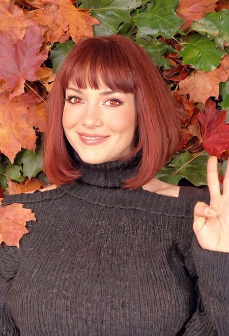 In the 2000s, candid photography, autumn, autumn leaves, black sweater, bob cut, breasts, brown coat, brown skirt, closed mouth, coat, large breasts, leaf, leaf background, long sleeves, looking at viewer, medium hair, off shoulder, orange background, red eyes, red hair, ribbed sweater, skirt, smile, solo, sweater, swept bangs, turtleneck, turtleneck sweater, upper body, waving, (soft lighting):1.2, film grain, sharp shadows, Samsung shot, atmospheric