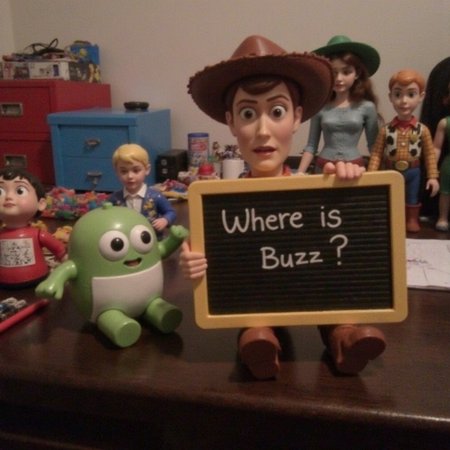 a photo of a toy figure of Wood from Toy Story, many different toys around, desk, child's room, desesperated expression, holding a toy writing board sign, writted "Where is buzz?", v8s, early 20's, muted and desaturated colors, low-lit, no humans around, only toys