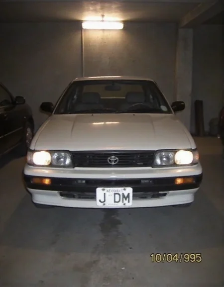 Toyota AE86, dusty, garaged, dim light from lamp above, headlights fogged up from wear, photo of front of car, license plate "JDM", date bottom right "10 04 1995",  <lora:2000analogcore_v3:1.5>