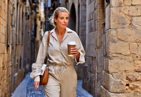 A hyper-detailed, high-resolution (8K) walking portrait of a 40-year-old woman (Like Catherine Deneuve) with sleek blonde hair tied back, walking through the Gothic Quarter of Barcelona with a takeaway coffee in hand. She wears a light beige jumpsuit with rolled-up sleeves, white slip-on shoes, and a crossbody leather satchel. A compact digital camera peeks out of the bag. Her natural beauty, including fine wrinkles and clear blue eyes, glows in the filtered Mediterranean light. Shot with Nikon D5300, 50mm lens, f/2.8, ISO 100. Background: blurred ancient stone arches and narrow cobbled streets. Photographic style: soft, clean, timeless — inspired by travel editorials.