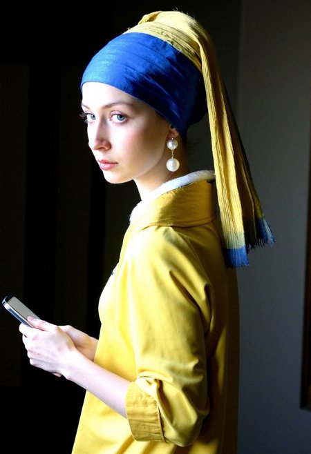 yellow dress, legs, holding iphone in her hand, young woman inspired by 'Girl with a Pearl Earring,' standing in a low-lit, dimly lit room. She wears a blue headscarf with golden fabric draped over it and a pearl earring that catches the subtle light, enhancing her classic, mysterious allure. Her gaze is directed over her shoulder, her soft, expressive eyes and delicate features highlighted by the ambient shadows in the room. room\u2019s interior. amateur quality with slight overexposure and grain. The low, moody lighting creates natural shadows that emphasize the texture of her attire and the contours of her face, enhancing the quiet, intimate atmosphere