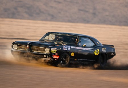 Create an ultra-detailed and realistic image of a race between 2 custom cars, created with parts from many muscle cars, with aerodynamic lines from 1972. The cars have tinted black windows. It is captured in motion with motion blur at full speed through a desert, daylight. Race between 2 cars. The photograph is captured with an Arri Alexa camera, with a zoom lens. <lora:Vintage Muscle Car SDXL:1>