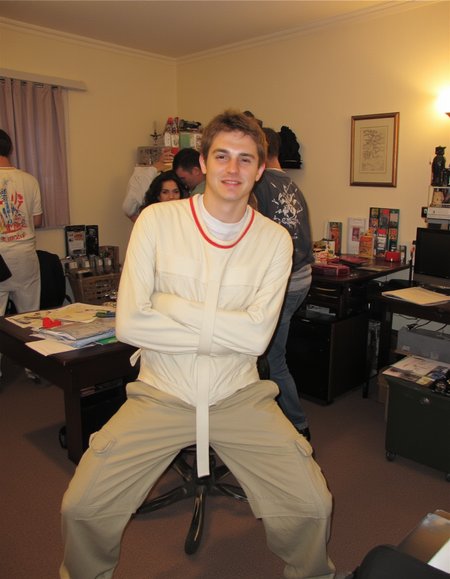 This is a photograph taken indoors, likely in an office or study room, showing a handsome, attractive young man in a playful, exaggerated pose. He is wearing a white sjposey and cargo pants. The man has short brown hair, with a playful, evil expression and smirk on his face. He is facing the left side, looking away from the camera. In the background, there are people, suggesting a house party. The image is a low quality old photo shot on old camera. The room has beige walls and a carpeted floor <lora:straitjacketflux-posey:1>  <lora:amateurphoto-6version:1>