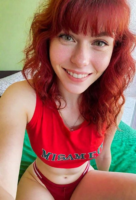 A full-body "Selfie taken with the front camera of an iPhone 15 in a close-up shot." female figure, a smiling and sexy redhead with bangs and wavy hair, dressed in a red and red t-shirt, and green shorts, sitting on a bed, looking at the camera, you can see her perfect breasts. , captured with a blurred background in an elegant hotel room. in leonard style, there is a natural light source illuminating the interior from a window, the background is blurred, the second part in hyper realistic style is made in a special test, by alex1shved, very realistic, rosy skin, beautiful lips and fleshy, smile, feeling of lightness and joy, hyperrealism, highly elaborate skin, direct gaze, by alex1shved