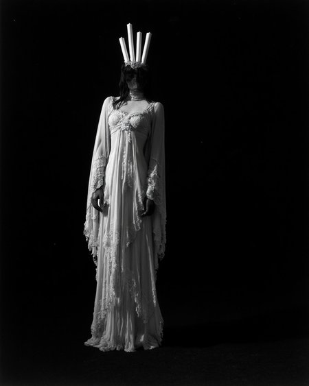 Kaos, The image is a black and white photograph of a woman standing against a stark black background, creating a dramatic, high-contrast scene. The woman is dressed in an elaborate, Gothic-inspired costume that resembles a Victorian-era mourning outfit, but with a surreal, theatrical twist. She is draped in a flowing, sheer, white fabric that cascades down to the floor, creating a ghostly, ethereal effect. The fabric is adorned with intricate lace patterns and delicate fringes, adding to the ethereal quality of her attire.
Her head is adorned with a headdress that features five tall, white candles arranged in a line above her head, creating a striking silhouette against the black background.