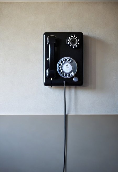 A stark black and white photograph of a vintage, wall-mounted rotary phone. The phone features a rectangular base affixed to a textured wall, with rotary dial. The handset hangs to the up, suspended by its coiled cord. The image is high-contrast, emphasizing the geometric shapes of the phone and the rough texture of the wall.