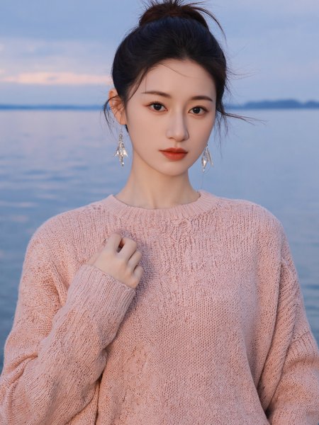 This photograph captures a young Asian woman standing by a serene, blue-hued lake with a distant, cloudy horizon. She is positioned slightly off-center to the right, adhering to the rule of thirds, creating a balanced composition. The woman has a slender physique, fair skin, and a delicate, refined face with subtle makeup. Her hair is styled in a casual bun with a few loose strands framing her face. She wears a light pink, textured knit sweater that adds a cozy, autumnal feel to the image. Her outfit is complemented by elegant, dangling silver earrings that catch the light, adding a touch of sophistication.

The background features a tranquil lake with gentle ripples, reflecting the soft, diffused light of either dawn or dusk, which casts a gentle glow on the scene. The sky is overcast, with a gradient of soft blues and grays, enhancing the calm and reflective mood of the image. The camera angle is at eye level, providing a clear, unobstructed view of the subject and the landscape. The overall lighting is natural and soft, with no harsh shadows, contributing to the serene and peaceful atmosphere.
