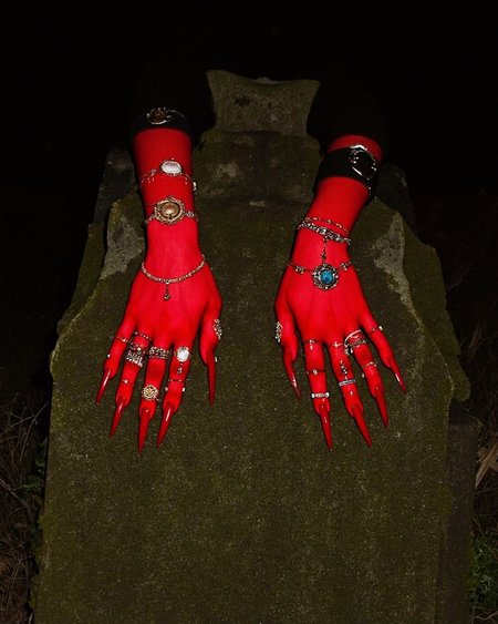 Kaos, The photograph depicts a scene at night, featuring a person with their hands positioned dramatically on an old, weathered gravestone. The person's skin is painted a vibrant red, which contrasts starkly with the grey, moss-covered stone. The hands are adorned with multiple rings, including a large, ornate piece on the middle finger of the left hand and smaller, more delicate rings on the other fingers. The fingernails are long and pointed, painted the same bright red as the skin. 
The hands are positioned on the stone, with the fingers splayed out, giving a sense of dramatic flair.