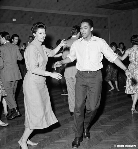 The picture is a black and white photograph depicting a lively dance scene, likely at a social event or club in the mid-20th century.  The focus is on a young Afro-American couple dancing. The woman, wearing a simple, elegant sweater and a knee-length skirt, is moving with grace. Her partner is stylishly dressed in a light-colored shirt and dark trousers. He appears to be leading the dance.  In the background, other couples and people are visible, enjoying the music. A saxophone player is noticeable amidst the dancers, indicating a jazz or swing atmosphere. The background is blurred, drawing attention to the main dancing couple in the foreground. The overall style suggests candid photography, capturing the spontaneous energy of the moment. The photographic style is reminiscent of photojournalism or street photography from the mid-20th century. The use of black and white enhances the contrast and mood, creating a timeless quality. The slightly grainy texture and focus on capturing movement in a realistic manner further contributes to the authenticity of the scene. The shallow depth of field keeps the focus sharply on the central dancing couple while maintaining enough context through the slightly blurred background.  The composition, with the central couple framed in the dance floor, guides the viewer's attention effectively. <lora:_Flux\Vintage_Jazz-Club.safetensors:1.0:1.0>