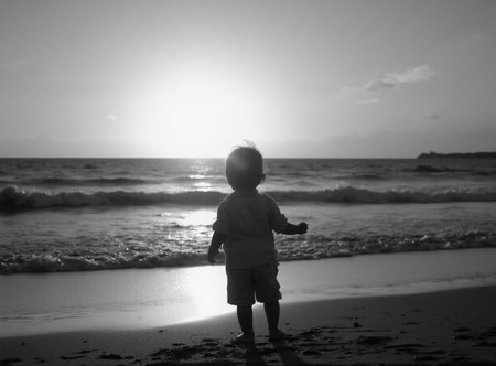 A black-and-white photograph of a child at the beach at sunset. The child is looking out toward the setting sun.
