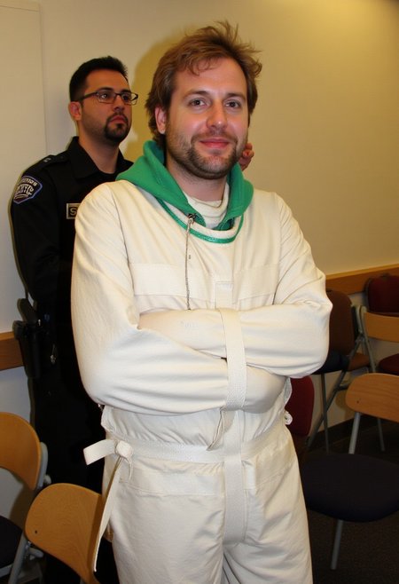 A photograph, likely in a lecture room. In the background, a man with short black hair and a beard, wearing a security guard uniform and glasses, is standing and doing something behind second person. The second person, a middle aged man with light brown wavy hair, untidy beard and earring is wearing a white sjposey, with a green hood behind his neck. He has a slight smile, suggesting a nervous mood. The room features a mix of wooden and upholstered chairs, adding to the casual atmosphere. <lora:straitjacketflux-posey:1.1>